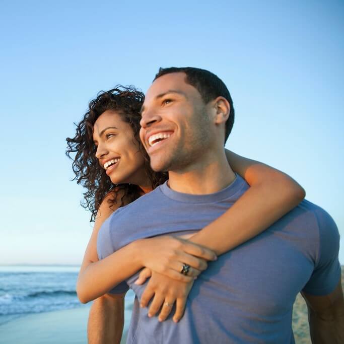 Smiling woman standing behind a man on the beach with her hands wrapped around his chest.