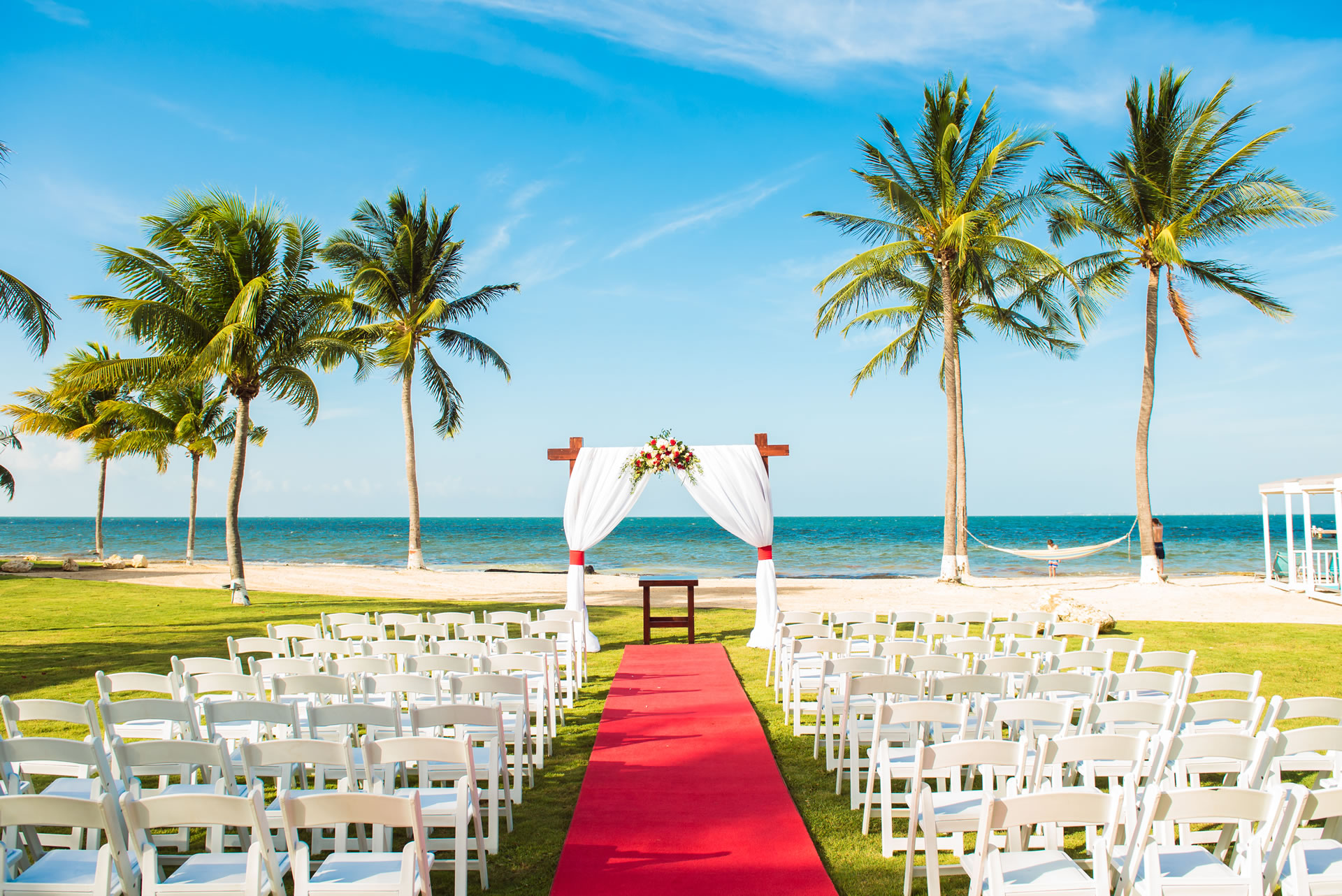 A wedding arch at the end of a red carpet dividing rows of white folding chairs. The beach of the Grand Caymanian Resort is in the background.