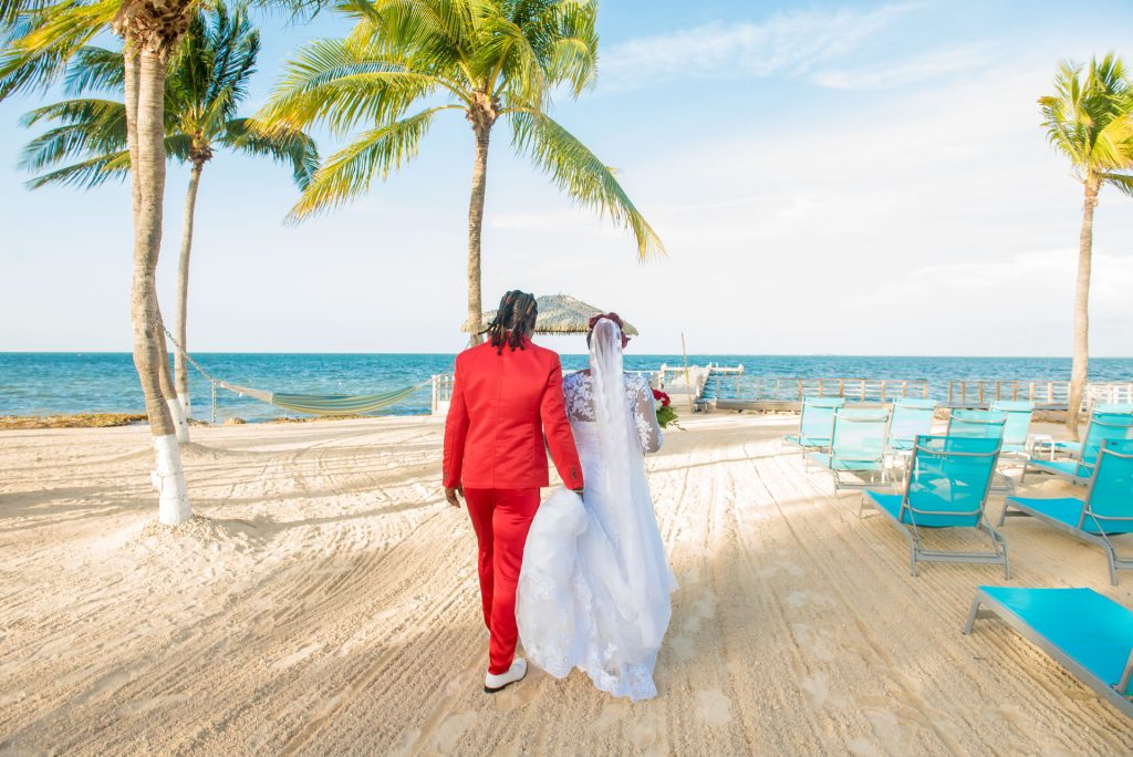 A man in a red suit escorts his bride in her bridal dress across the beach towards the fishing pier.