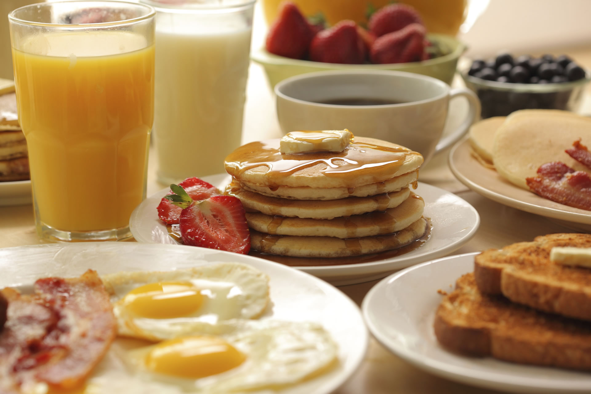 Breakfast spread featuring pancakes and maple syrup, sunny-side up eggs, bacon, toast, orange juice, milk, coffee, and berries.