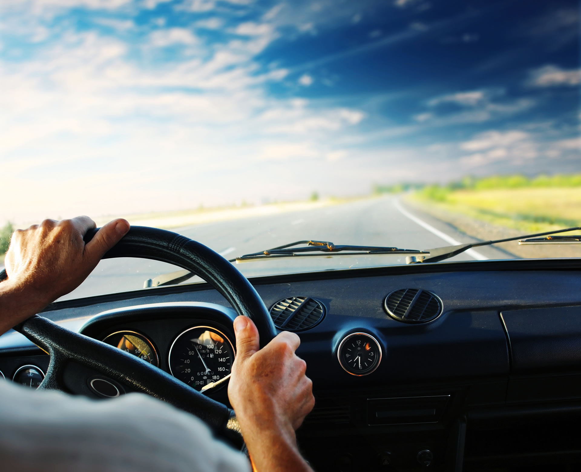 Close up of the a man gripping a steering wheel and the dashboard of his car as he drives down an open road.