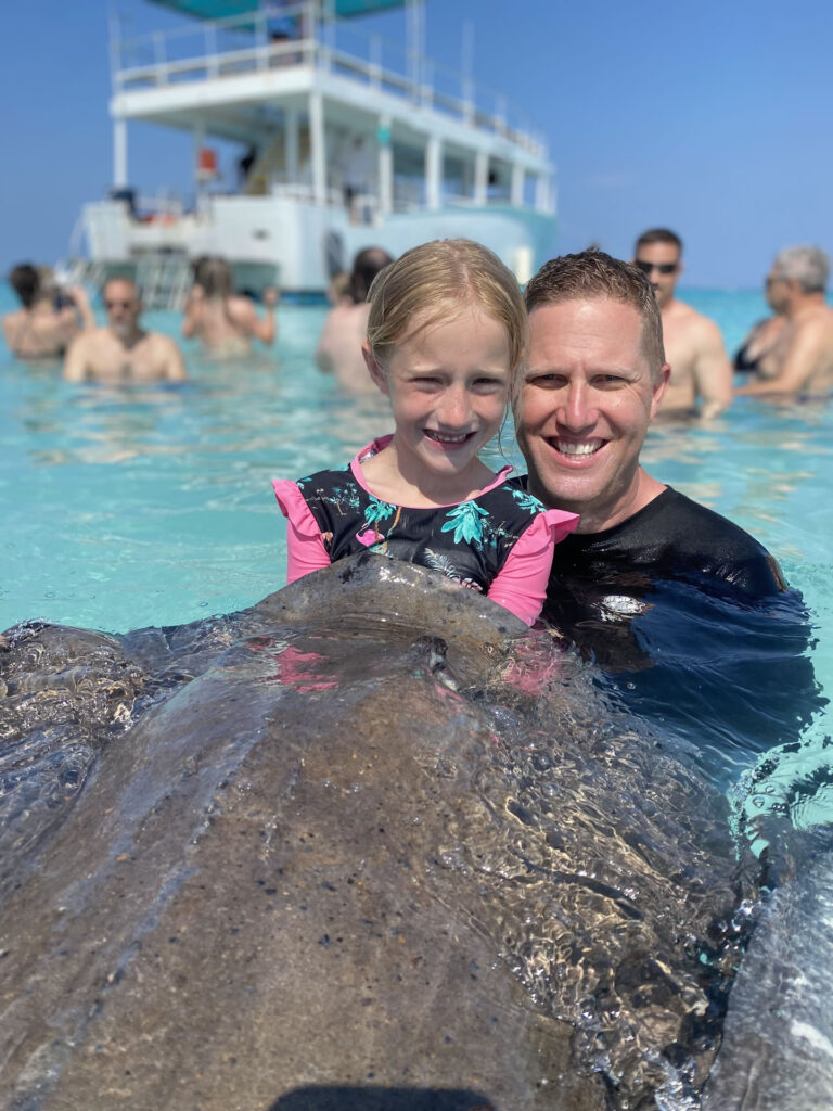 A dad and his daughter in the ocean next to a stingray on the Cayman Sun Splash Tour.