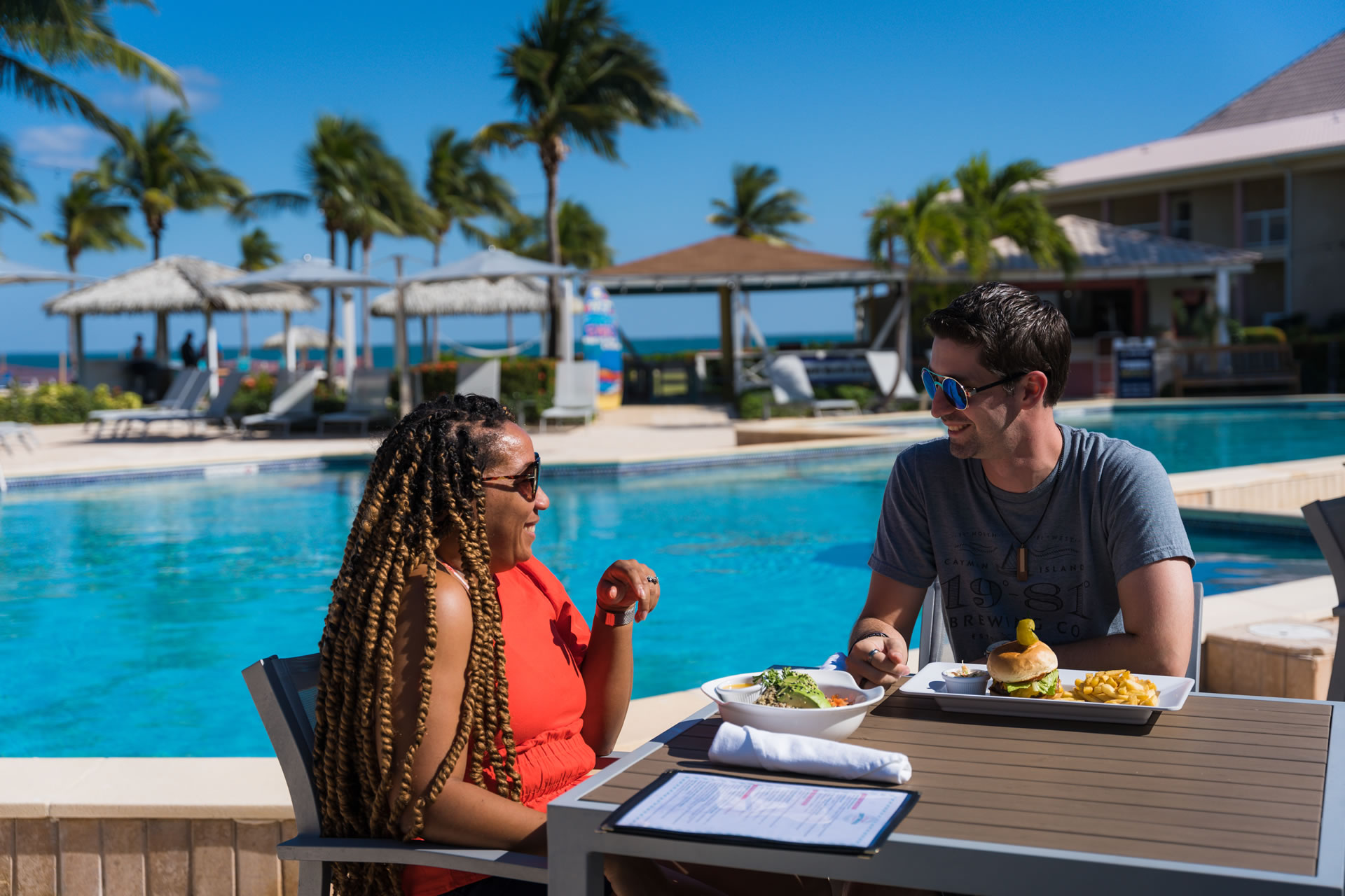 A man and woman dining near the pool of the Grand Caymanian Resort on a sunny day.