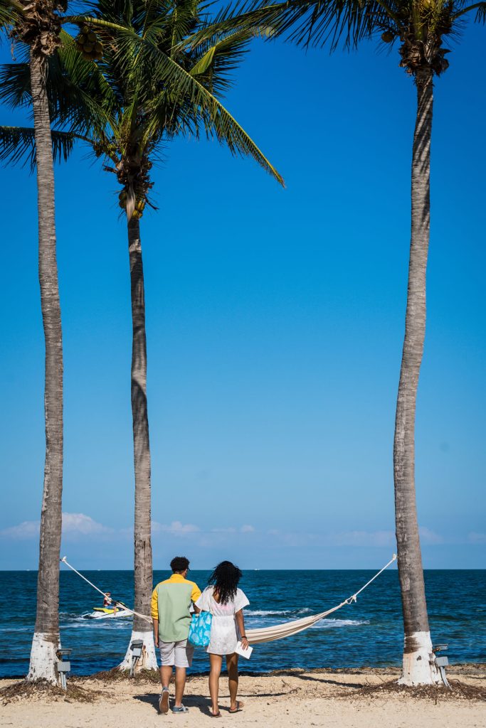 A couple walking towards a hammock strung up between two palm trees on the beach at the Grand Caymanian Resort.