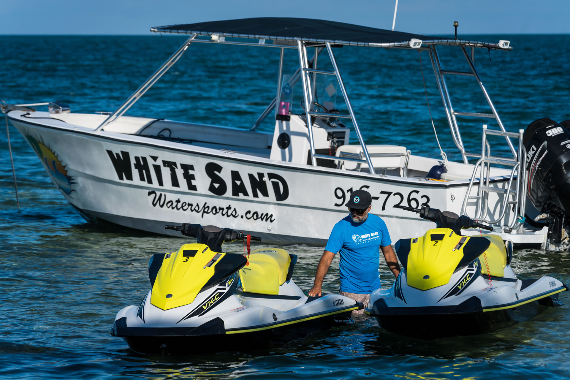 A man standing in the water next to two personal watercraft and a boat with the White Sand logo and watersports.com on the side.