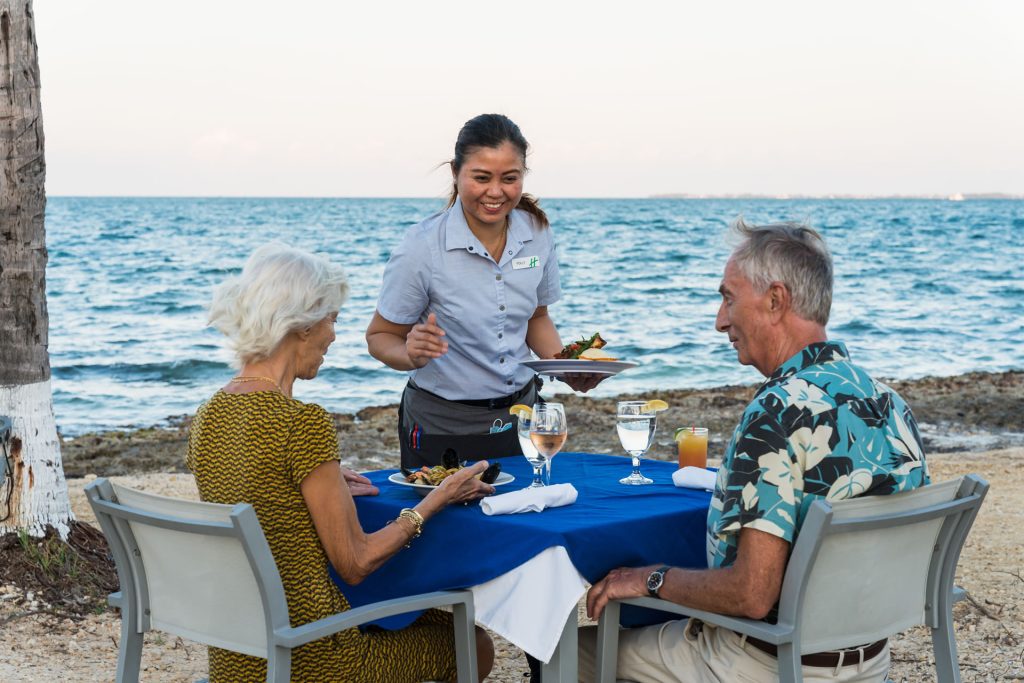 An elderly couple dining on the beach at the Grand Caymanian Resort. A server is bringing their food.